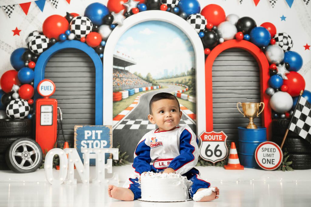 Baby Sitting Behind A White Birthday Cake During A Race Car Cake Smash Photoshoot In Dallas